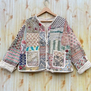May include: A cream-coloured quilted jacket with a patchwork design featuring various floral and geometric patterns in shades of pink, blue, green, and brown. The jacket has a button closure and two patch pockets.