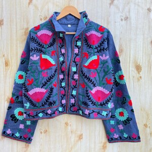 K&ouml;nnte beinhalten: Eine blaue bestickte Jacke mit Blumenmustern in den Farben Rosa, Rot, Gr&uuml;n und Schwarz. Die Jacke hat einen Stehkragen und einen Rei&szlig;verschluss vorne.