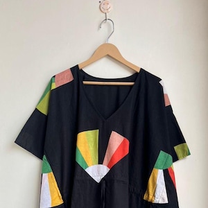 May include: Black caftan dress with a V-neck and short sleeves. The dress features colorful fan-shaped patterns in yellow, green, red, and white. A drawstring cinches the waist. The dress is hanging on a wooden hanger.
