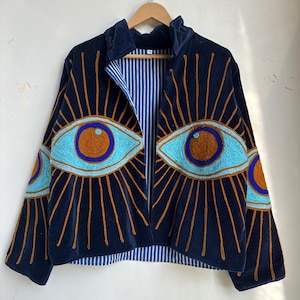 May include: Navy blue velvet jacket with embroidered eye designs in turquoise and brown. The jacket features a striped blue and white lining and a zip-up front. The eye designs are on the front and sleeves.