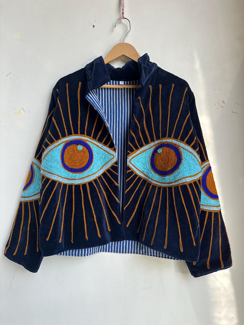 May include: Navy blue velvet jacket with embroidered eye designs in turquoise and brown. The jacket features a striped blue and white lining and a zip-up front. The eye designs are on the front and sleeves.