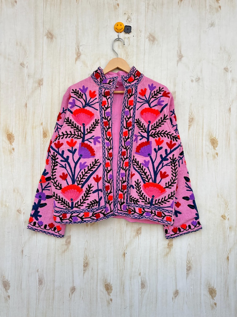 May include: A pink embroidered jacket with floral designs in red, blue, and purple. The jacket has a mandarin collar and long sleeves. The jacket is hanging on a hanger against a light wood background.