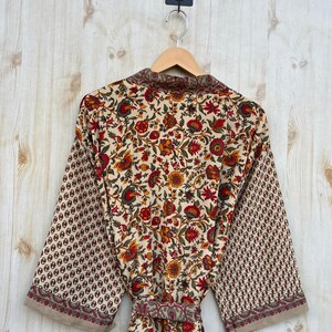 May include: A floral print robe with a cream background, red, yellow, and green flowers, and brown accents. The sleeves have a repeating pattern of small brown circles. The robe is hanging on a wooden hanger.