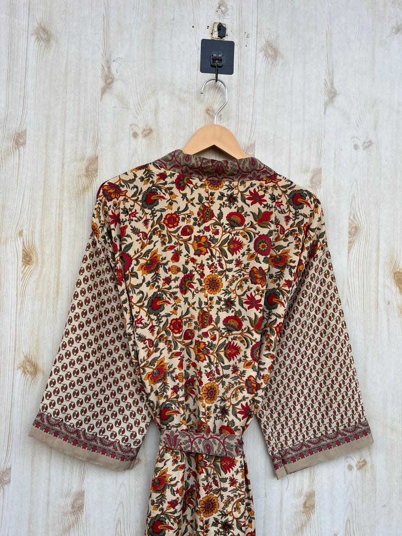 May include: A floral print robe with a cream background, red, yellow, and green flowers, and brown accents. The sleeves have a repeating pattern of small brown circles. The robe is hanging on a wooden hanger.