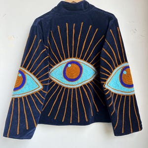 May include: Navy blue velvet jacket with embroidered eye designs on the sleeves and back. The eyes are light blue with brown and purple accents, surrounded by gold rays. The jacket has a high collar.