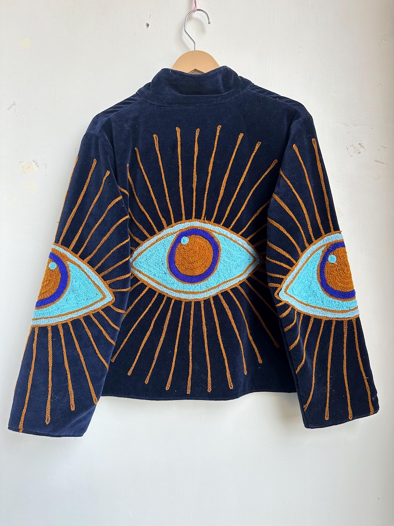 May include: Navy blue velvet jacket with embroidered eye designs on the sleeves and back. The eyes are light blue with brown and purple accents, surrounded by gold rays. The jacket has a high collar.
