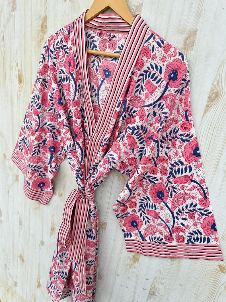 K&ouml;nnte beinhalten: Ein Bademantel mit Blumenmuster in Rosa, Wei&szlig; und Marineblau h&auml;ngt an einem Holzb&uuml;gel. Der Bademantel zeigt ein sich wiederholendes Muster aus gro&szlig;en rosa Blumen und blauen Bl&auml;ttern auf wei&szlig;em Grund. Er hat eine gestreifte Bord&uuml;re und eine passende Krawatte.