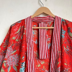 Pu&ograve; includere: Un accappatoio rosso in stile kimono con un motivo floreale e di uccelli. L'accappatoio presenta strisce verticali rosse e bianche lungo i bordi e il colletto. L'accappatoio &egrave; appeso a una gruccia di legno.