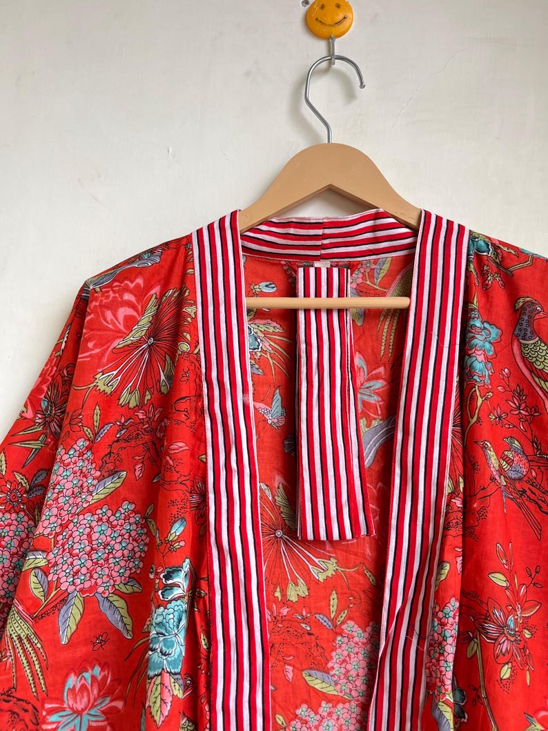 Pu&ograve; includere: Un accappatoio rosso in stile kimono con un motivo floreale e di uccelli. L'accappatoio presenta strisce verticali rosse e bianche lungo i bordi e il colletto. L'accappatoio &egrave; appeso a una gruccia di legno.