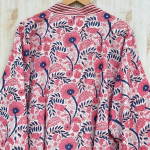 K&ouml;nnte beinhalten: Ein wei&szlig;er Kimono-Bademantel mit einem lebendigen Blumenmuster. Das Design zeigt gro&szlig;e rosa Bl&uuml;ten mit blauen Zentren und dunkelblauen Bl&auml;ttern. Der Bademantel hat eine gestreifte Bord&uuml;re und h&auml;ngt an einem Holzb&uuml;gel.
