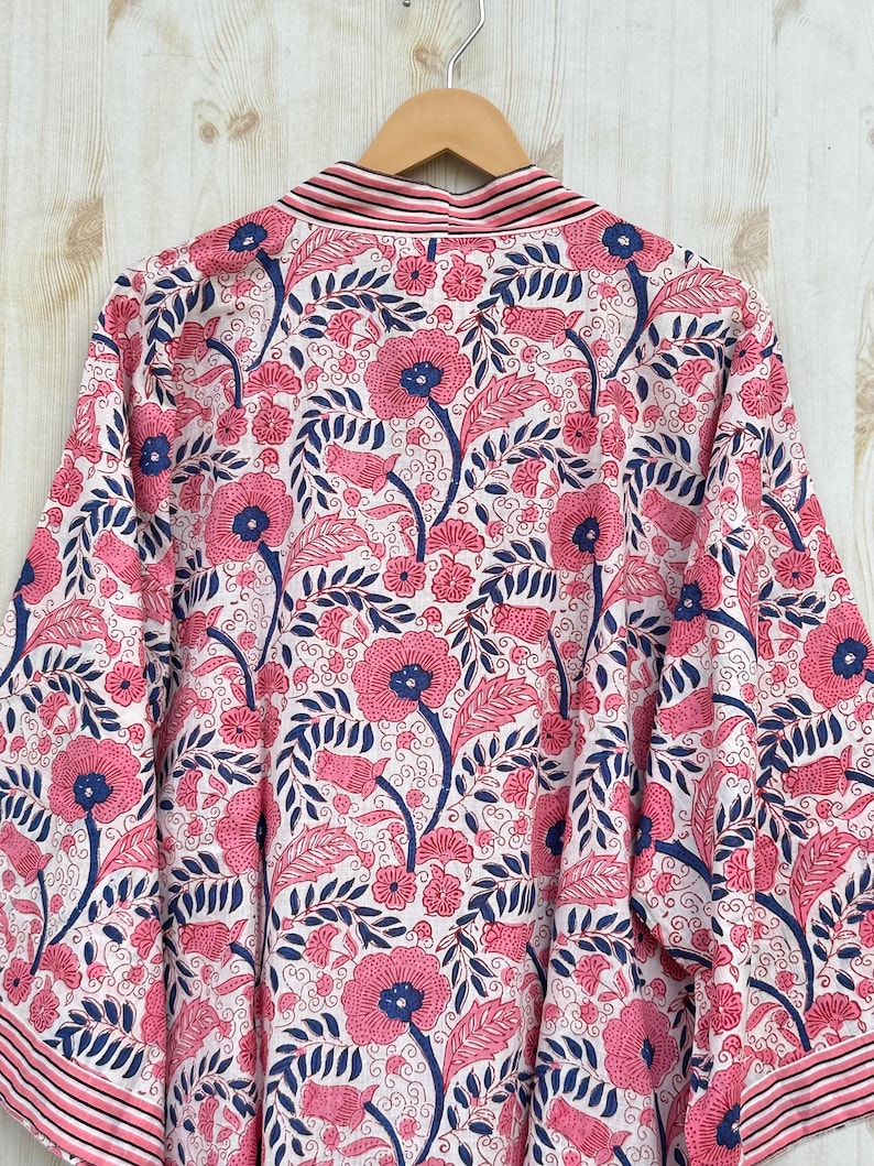 K&ouml;nnte beinhalten: Ein wei&szlig;er Kimono-Bademantel mit einem lebendigen Blumenmuster. Das Design zeigt gro&szlig;e rosa Bl&uuml;ten mit blauen Zentren und dunkelblauen Bl&auml;ttern. Der Bademantel hat eine gestreifte Bord&uuml;re und h&auml;ngt an einem Holzb&uuml;gel.