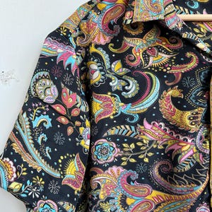 Op de afbeelding: Zwart overhemd met korte mouwen met een levendig paisley-patroon in geel, roze, blauw en groen. Het overhemd heeft een klassieke kraag en hangt aan een houten hanger. Het maatlabel luidt "M".