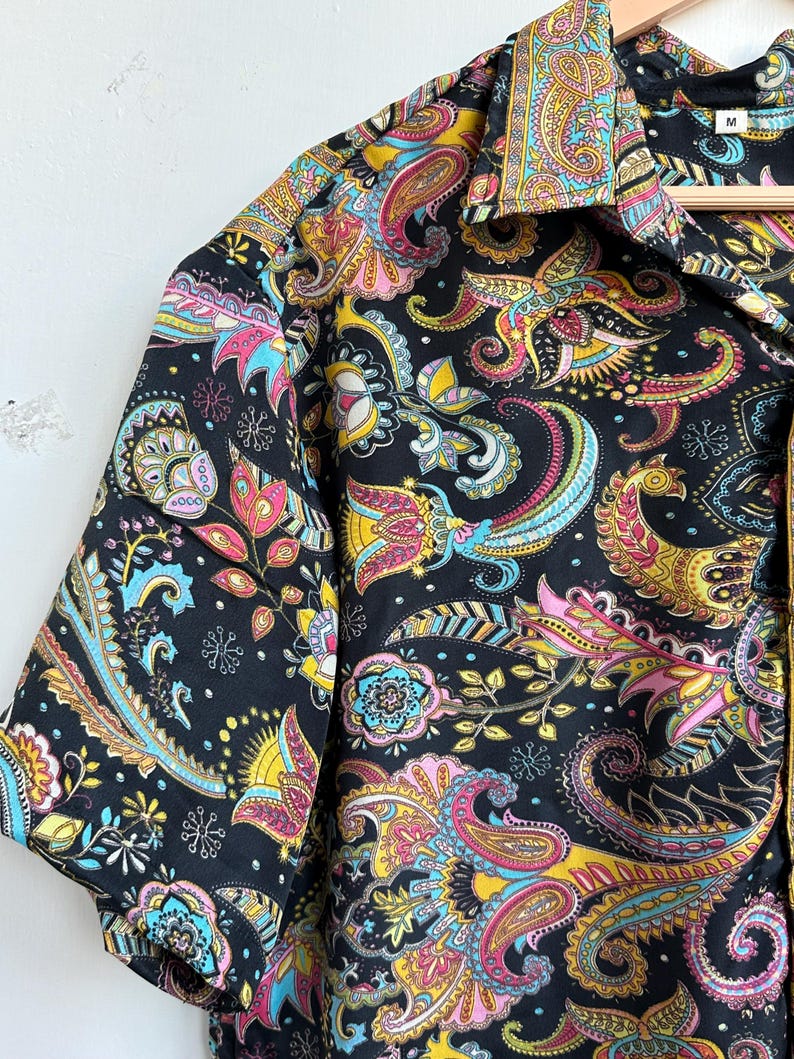 Op de afbeelding: Zwart overhemd met korte mouwen met een levendig paisley-patroon in geel, roze, blauw en groen. Het overhemd heeft een klassieke kraag en hangt aan een houten hanger. Het maatlabel luidt "M".