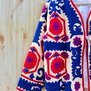 May include: Cream-coloured embroidered jacket with a floral pattern in red, blue, and purple. The jacket has a V-neck and long sleeves. The embroidery is detailed and covers the front and sleeves.