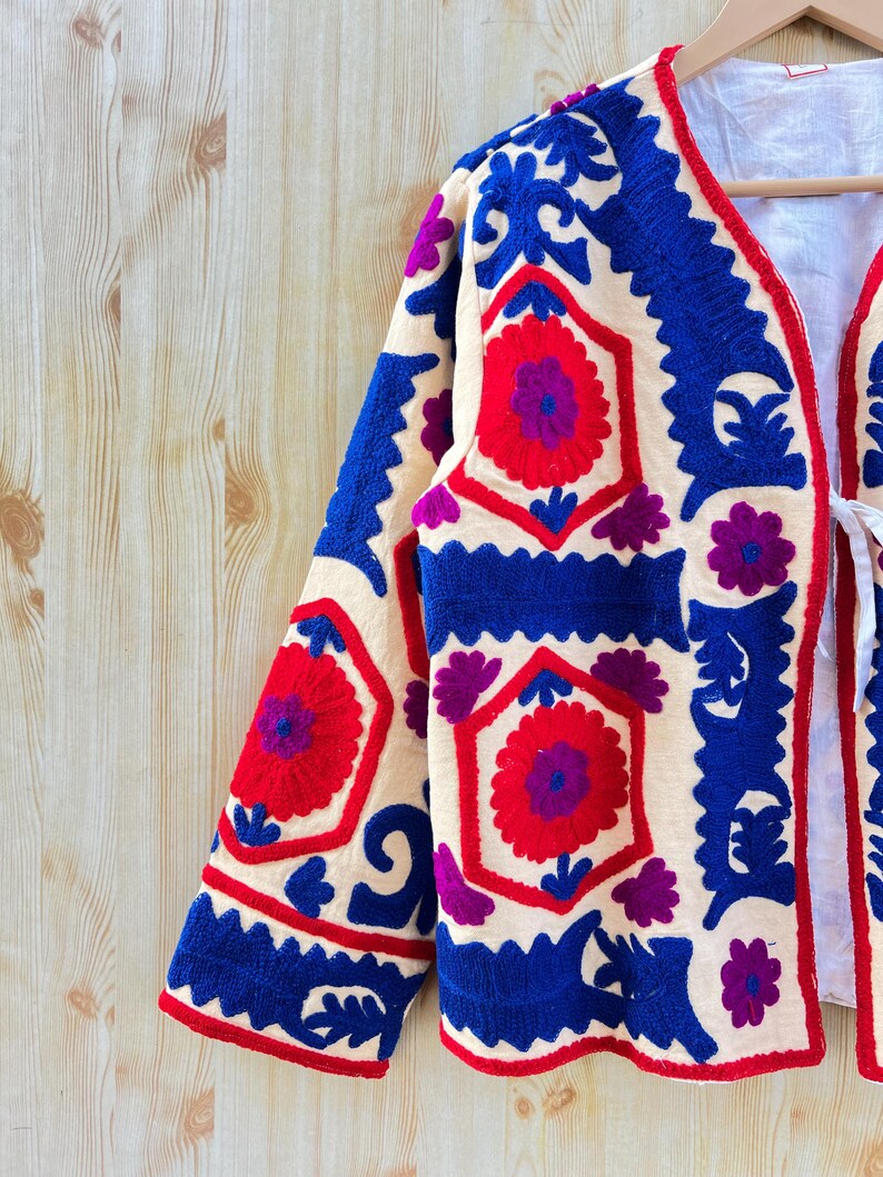 May include: Cream-coloured embroidered jacket with a floral pattern in red, blue, and purple. The jacket has a V-neck and long sleeves. The embroidery is detailed and covers the front and sleeves.