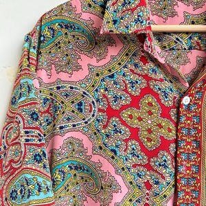 Op de afbeelding: Een shirt met korte mouwen met een levendig paisley-patroon in roze, rood, geel en blauwgroen. Het shirt heeft een kraag en is aan de voorkant dichtgeknoopt. Het maatlabel luidt "M".