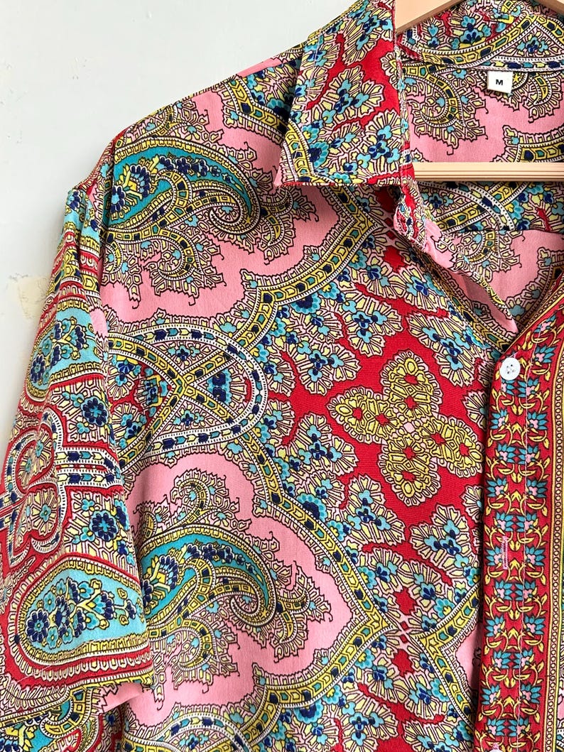 Op de afbeelding: Een shirt met korte mouwen met een levendig paisley-patroon in roze, rood, geel en blauwgroen. Het shirt heeft een kraag en is aan de voorkant dichtgeknoopt. Het maatlabel luidt "M".