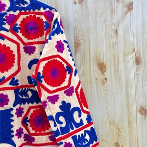 May include: Cream-coloured jacket with detailed embroidery in red, blue, and purple. The design features floral and geometric patterns. The jacket's sleeves are visible, showcasing the detailed stitching and vibrant colour palette.