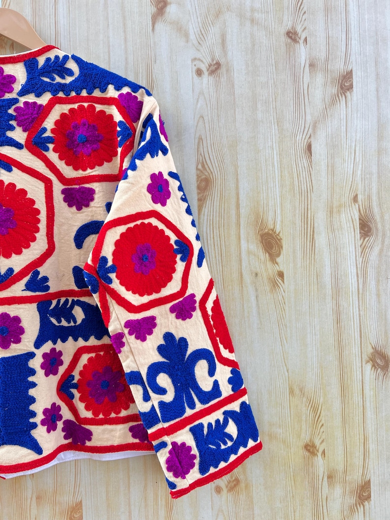 May include: Cream-coloured jacket with detailed embroidery in red, blue, and purple. The design features floral and geometric patterns. The jacket's sleeves are visible, showcasing the detailed stitching and vibrant colour palette.
