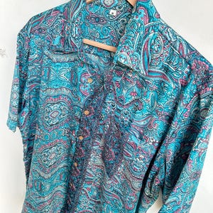 Op de afbeelding: Een overhemd met korte mouwen met een levendig paisley-patroon in tinten turquoise, roze en wit. Het overhemd hangt aan een houten hanger. De stof heeft een zijdeachtige glans en het ontwerp is ingewikkeld en gedetailleerd.