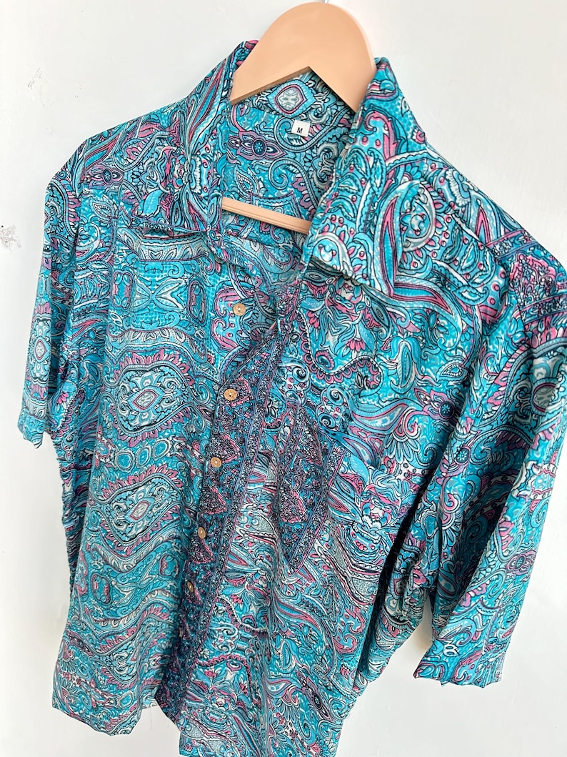 Op de afbeelding: Een overhemd met korte mouwen met een levendig paisley-patroon in tinten turquoise, roze en wit. Het overhemd hangt aan een houten hanger. De stof heeft een zijdeachtige glans en het ontwerp is ingewikkeld en gedetailleerd.