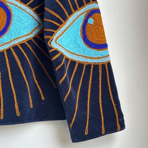 May include: Navy blue jacket with embroidered eye designs. The eyes are light blue with brown and blue accents, surrounded by gold rays. The jacket's fabric is a dark blue, and the embroidery adds texture and detail.