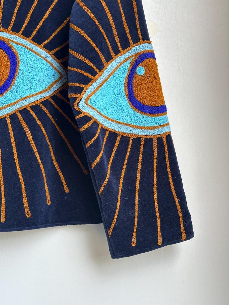 May include: Navy blue jacket with embroidered eye designs. The eyes are light blue with brown and blue accents, surrounded by gold rays. The jacket's fabric is a dark blue, and the embroidery adds texture and detail.