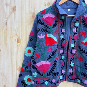 K&ouml;nnte beinhalten: Eine blaue bestickte Jacke mit Blumenmustern in den Farben Rosa, Rot, Gr&uuml;n und Schwarz. Die Jacke hat einen Rei&szlig;verschluss und einen Stehkragen.
