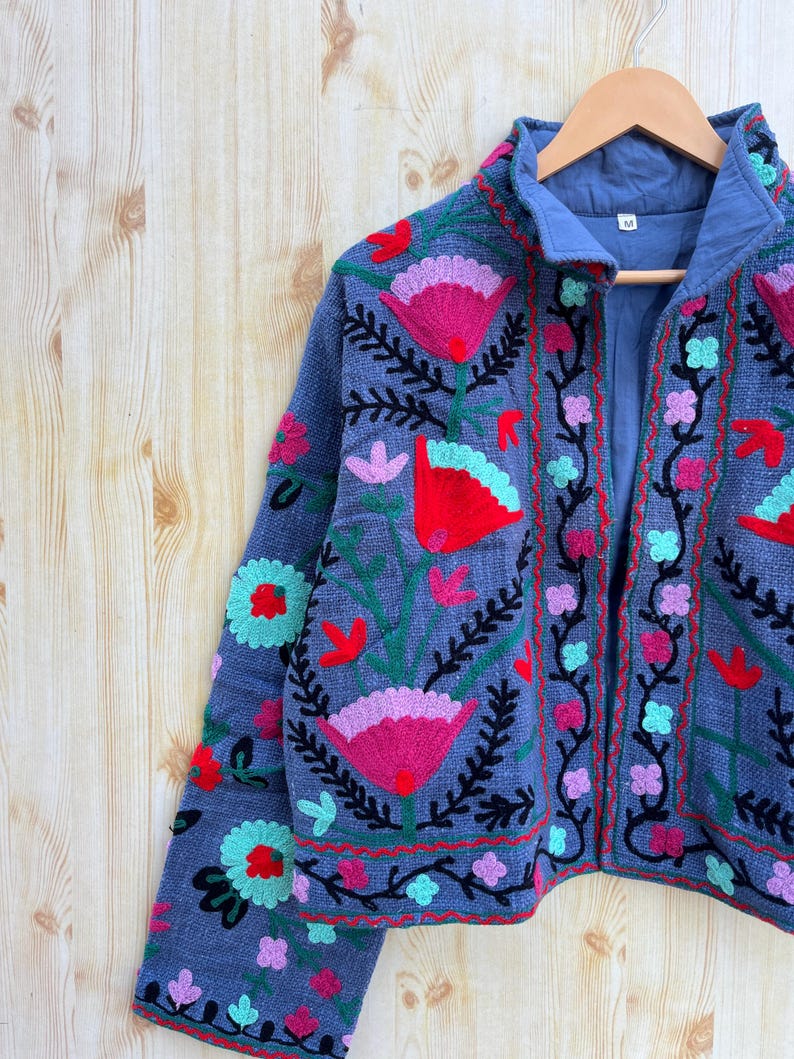 K&ouml;nnte beinhalten: Eine blaue bestickte Jacke mit Blumenmustern in den Farben Rosa, Rot, Gr&uuml;n und Schwarz. Die Jacke hat einen Rei&szlig;verschluss und einen Stehkragen.