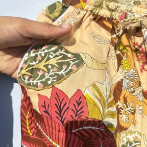 Op de afbeelding: Beige pyjamabroek met een bloemenprint met rode, groene en gele bladeren. De broek heeft een witte elastische tailleband en een trekkoord.