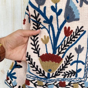 Pode incluir: Close-up de um casaco bege com bordados florais coloridos. O casaco apresenta um design detalhado com flores e folhas bordadas em azul, vermelho, amarelo e preto. A borda do casaco tamb&eacute;m &eacute; decorada com um padr&atilde;o bordado semelhante.