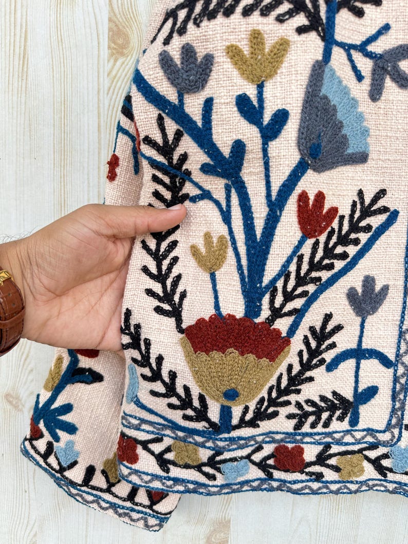 Pode incluir: Close-up de um casaco bege com bordados florais coloridos. O casaco apresenta um design detalhado com flores e folhas bordadas em azul, vermelho, amarelo e preto. A borda do casaco tamb&eacute;m &eacute; decorada com um padr&atilde;o bordado semelhante.