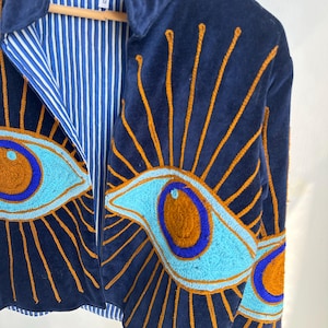 May include: Navy blue velvet jacket with embroidered eye designs in shades of blue and orange. The jacket features a collared neckline and a striped blue and white lining. The eye designs are on the front and sleeves.