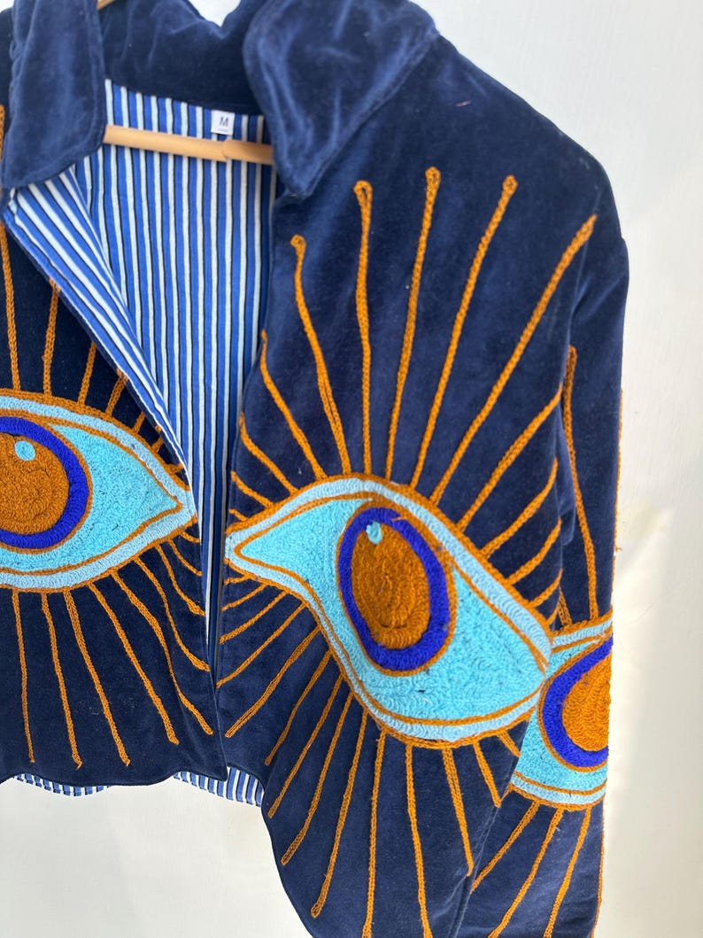 May include: Navy blue velvet jacket with embroidered eye designs in shades of blue and orange. The jacket features a collared neckline and a striped blue and white lining. The eye designs are on the front and sleeves.