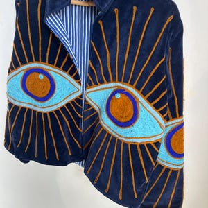 May include: Navy blue velvet jacket with embroidered eye design. The jacket features a light blue and brown eye pattern with gold sunburst rays. The interior lining is striped blue and white. The jacket is hanging on a wooden hanger.
