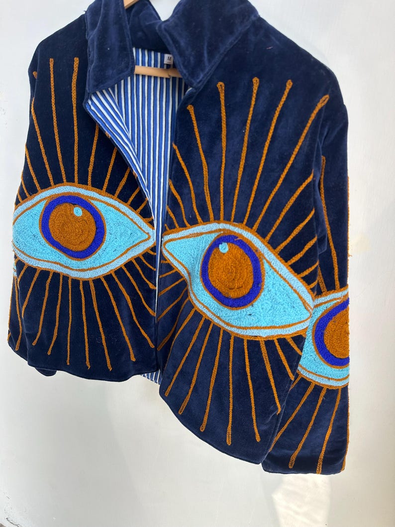 May include: Navy blue velvet jacket with embroidered eye design. The jacket features a light blue and brown eye pattern with gold sunburst rays. The interior lining is striped blue and white. The jacket is hanging on a wooden hanger.