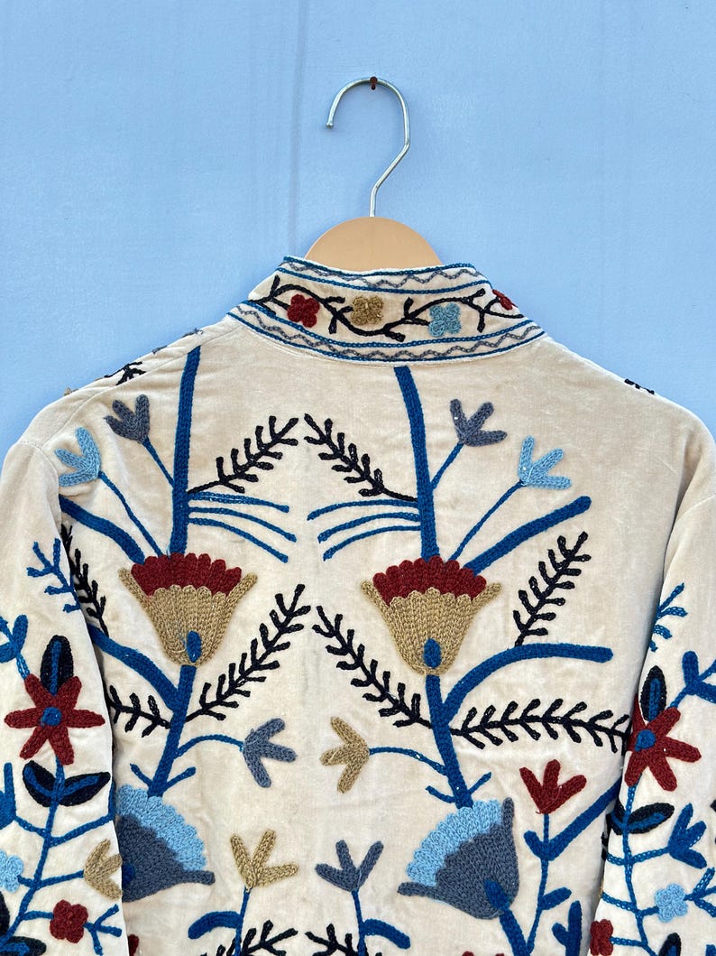 Pu&ograve; includere: Una giacca color crema con ricami floreali blu, rossi e dorati. La giacca ha un collo alto e maniche lunghe.