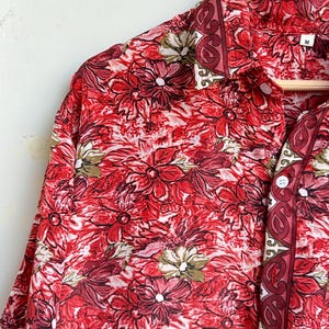 Op de afbeelding: Rood bloemen overhemd met een levendig patroon van rode en witte bloemen. Het overhemd heeft een kraag en korte mouwen. Het overhemd hangt aan een houten hanger.