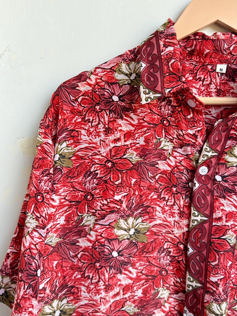 Op de afbeelding: Rood bloemen overhemd met een levendig patroon van rode en witte bloemen. Het overhemd heeft een kraag en korte mouwen. Het overhemd hangt aan een houten hanger.