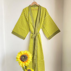 May include: A lime green and white checkered robe with olive green trim and a matching belt hangs on a wooden hanger. The robe is displayed against a white wall, with a sunflower in the foreground.
