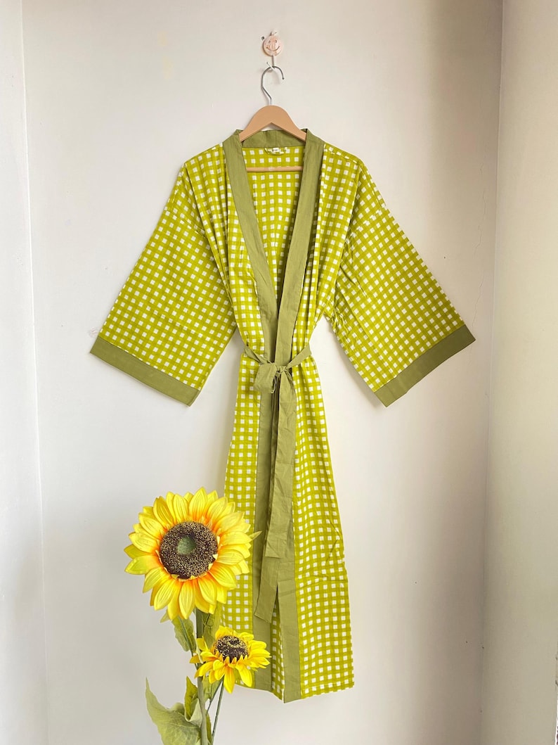 May include: A lime green and white checkered robe with olive green trim and a matching belt hangs on a wooden hanger. The robe is displayed against a white wall, with a sunflower in the foreground.