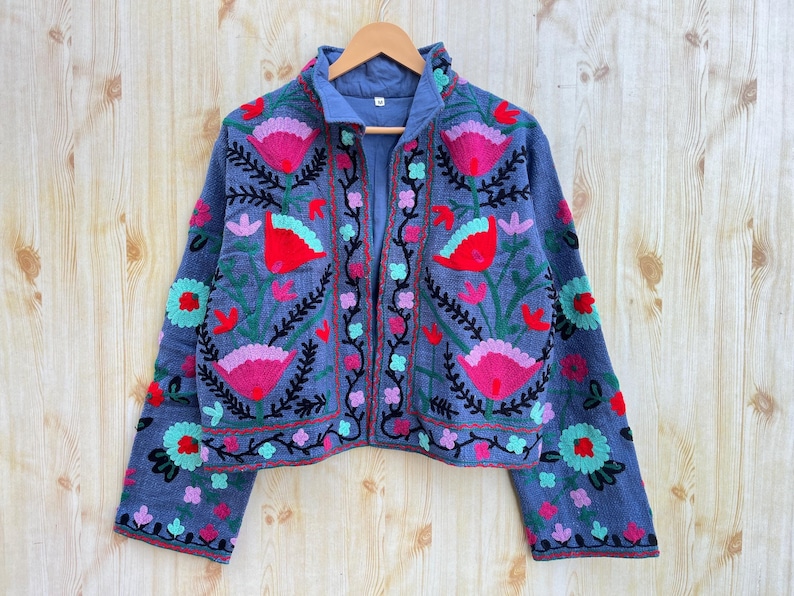 K&ouml;nnte beinhalten: Eine blaue bestickte Jacke mit Blumenmustern in den Farben Rosa, Rot, Gr&uuml;n und Schwarz. Die Jacke hat einen Stehkragen und einen Rei&szlig;verschluss vorne.