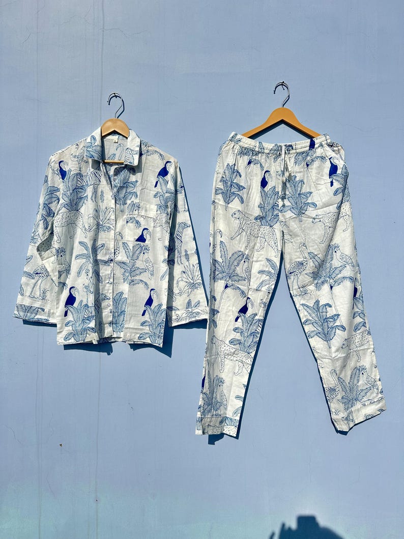 May include: A white pyjama set with a blue and white tropical print featuring toucans and palm trees. The set includes a long-sleeved shirt with a button-up front and matching trousers with an elastic waistband.