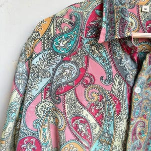 Op de afbeelding: Een kleurrijk paisley-patroonshirt in roze, turquoise, geel en wit. Het shirt hangt aan een houten hanger. De stof heeft een gedetailleerd ontwerp met gebogen, druppelvormige motieven.