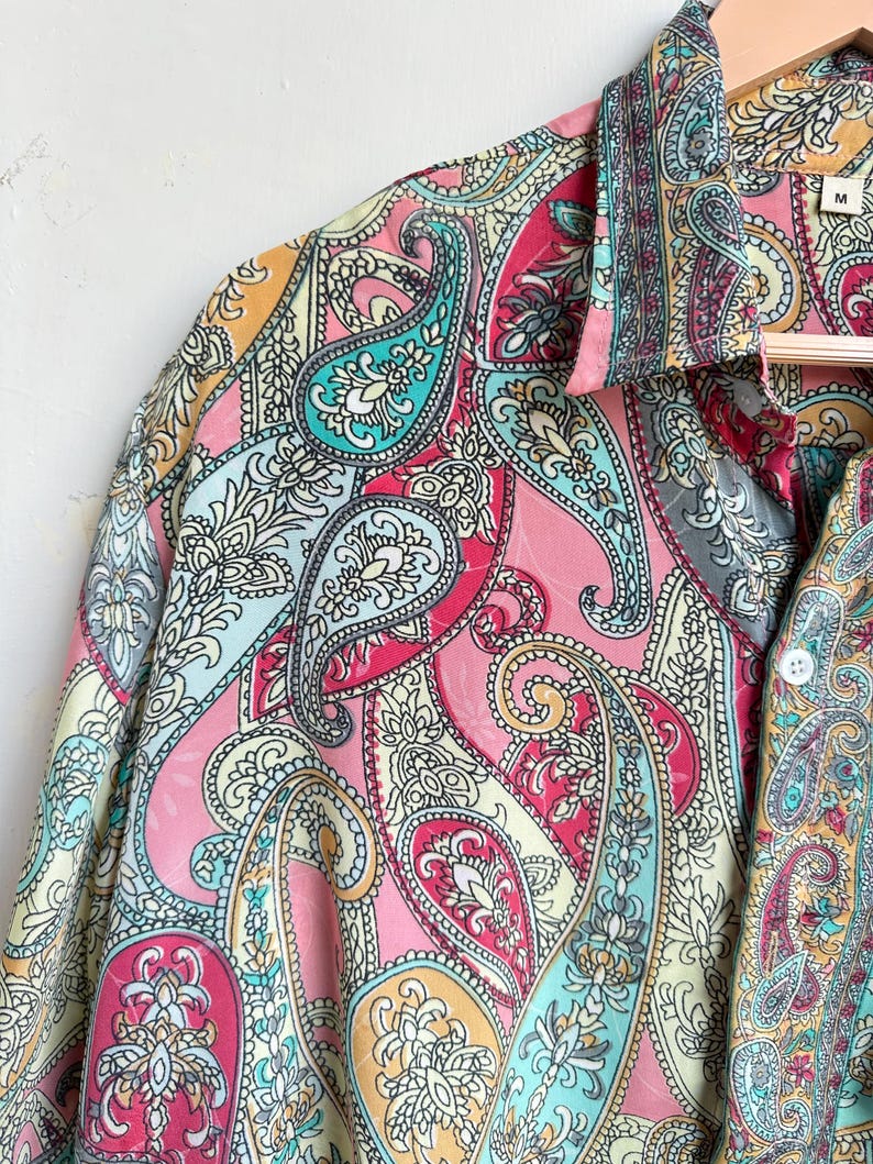 Op de afbeelding: Een kleurrijk paisley-patroonshirt in roze, turquoise, geel en wit. Het shirt hangt aan een houten hanger. De stof heeft een gedetailleerd ontwerp met gebogen, druppelvormige motieven.