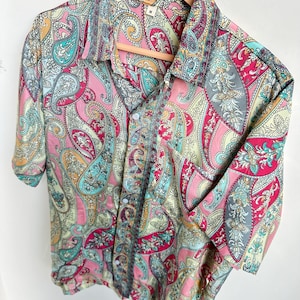 Op de afbeelding: Een overhemd met korte mouwen en een levendig paisley-patroon in roze, turquoise, geel en grijs. Het overhemd hangt aan een houten kleerhanger, wat het gedetailleerde ontwerp en kleurenpalet benadrukt.