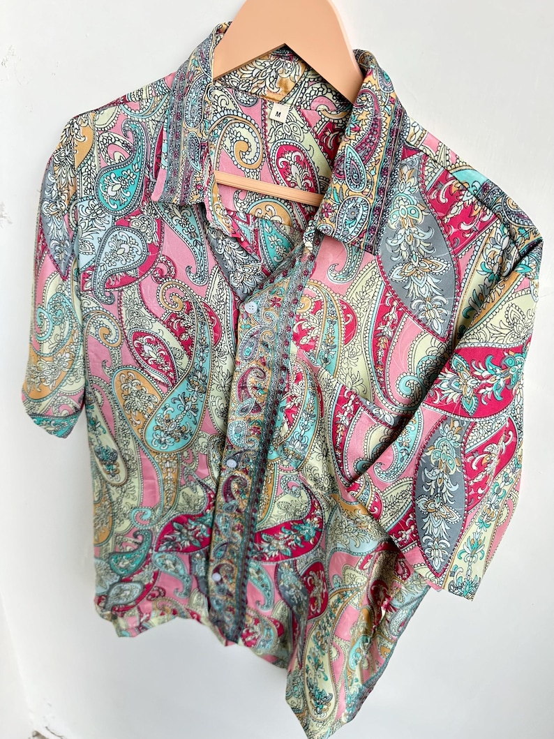 Op de afbeelding: Een overhemd met korte mouwen en een levendig paisley-patroon in roze, turquoise, geel en grijs. Het overhemd hangt aan een houten kleerhanger, wat het gedetailleerde ontwerp en kleurenpalet benadrukt.