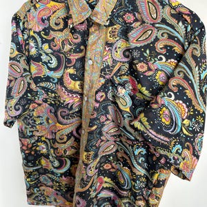 Op de afbeelding: Een zwart overhemd met knopen en een kleurrijk paisley patroon. Het paisley ontwerp bevat tinten geel, roze, blauw en groen. Het overhemd heeft een kraag en korte mouwen.