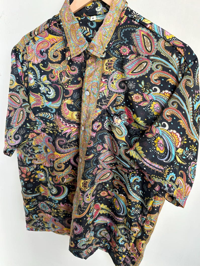 Op de afbeelding: Een zwart overhemd met knopen en een kleurrijk paisley patroon. Het paisley ontwerp bevat tinten geel, roze, blauw en groen. Het overhemd heeft een kraag en korte mouwen.