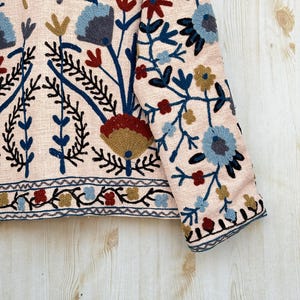 Pode incluir: Close-up de um casaco rosa claro com bordados florais intrincados. O design apresenta flores azuis, vermelhas e douradas com caules e folhas pretas. A bainha e os punhos do casaco t&ecirc;m uma borda detalhada.